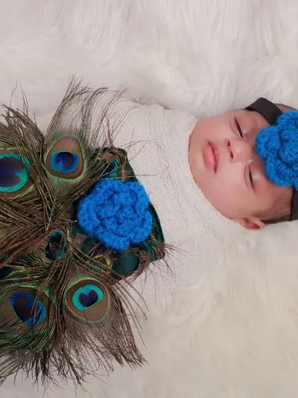 A beautiful overhead shot of a newborn decorated with peacock feathers, creating a stunning visual pattern. The blue knitted flowers add a lovely touch of color.