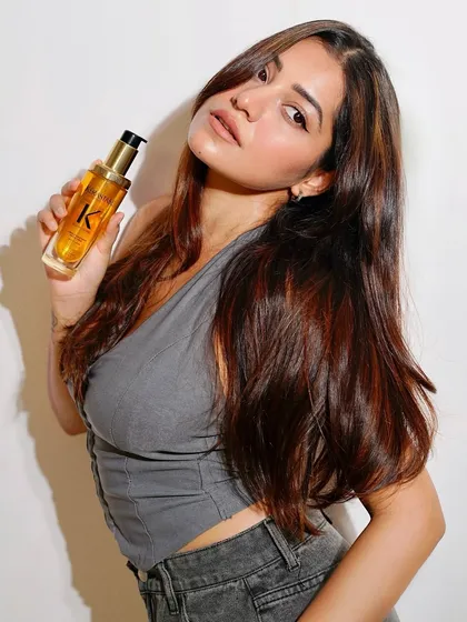 A model showcasing her radiant, healthy hair while holding a bottle of Kérastase Elixir Ultime, a luxury hair oil we use to provide ultimate shine and nourishment.