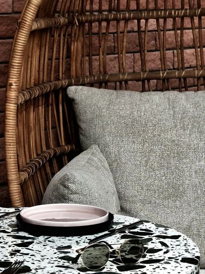 A detail shot capturing the textures of the space: the rough weave of the rattan chair, the smooth, speckled terrazzo tabletop, and the soft fabric of the cushion. Every material is chosen to delight the senses.