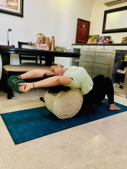 To open the shoulders for deeper backbends, we use props. Here, a student uses a bolster for support and light dumbbells to add gentle weight, increasing the stretch across the chest and front deltoids.