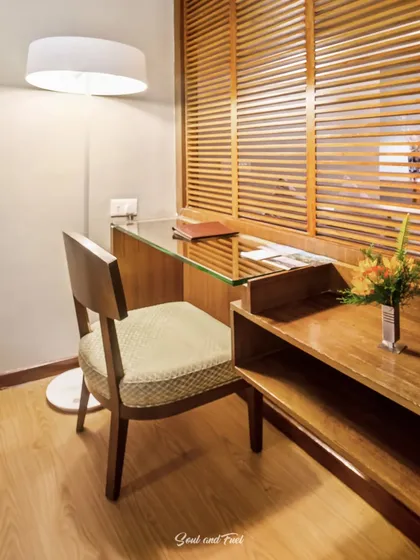 A functional and stylish wooden study desk in a hotel room. The slatted wood screen and floor lamp add a touch of modern design to the comfortable workspace.