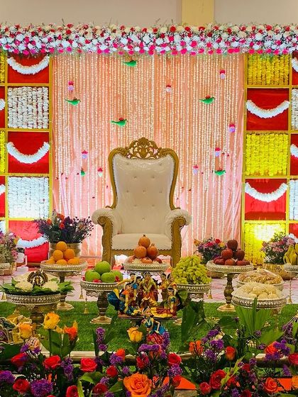 A colorful and festive Seemantham stage. The backdrop features red, yellow, and white panels with hanging garlands, and the foreground is filled with a beautiful array of flowers and offerings.
