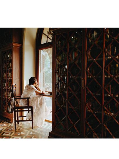 A moment of reflection by the window in a library filled with old books. The light and composition create a serene, literary mood for the 'Banaras, Bombay' campaign.
