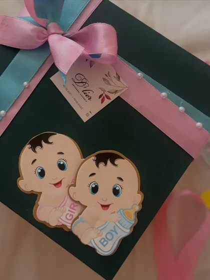 A close-up of the "mom-to-be" gift box, showing the detail of the pink and blue ribbons and the charming baby boy and girl cutouts.