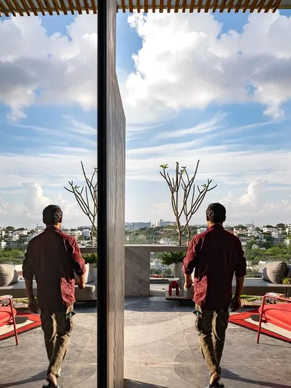 A figure walks across a reflective glass door on a rooftop terrace, with the open sky and cityscape mirrored in the surface. The design plays with light, reflection, and views to create a dynamic space.