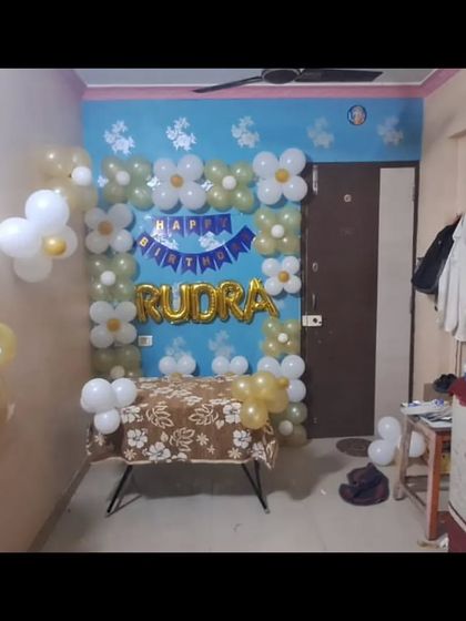 A simple birthday decoration at home with a 'Happy Birthday Rudra' foil balloon and a frame of white and gold balloons.