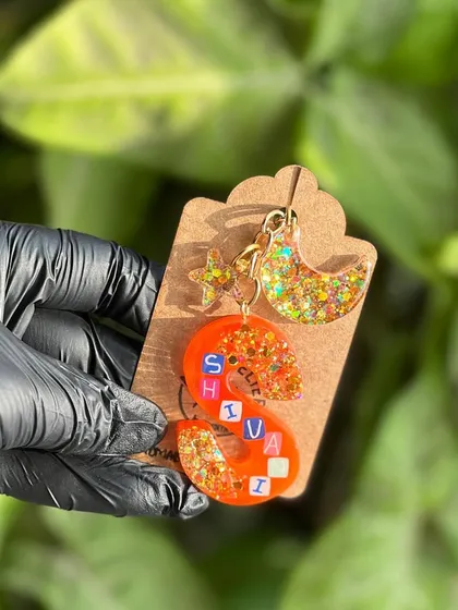 A custom 'Shivai' keychain with a moon and star charm. The combination of the orange resin, glitter, and personalized name makes it a truly unique accessory.