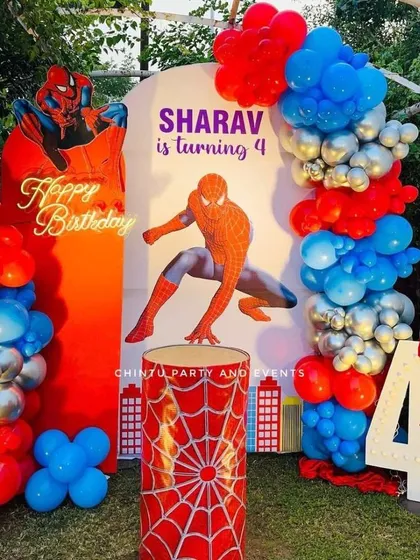 An action-packed Spiderman theme for a 4th birthday, perfect for an outdoor setting. The decoration includes Spiderman cutouts, a city-themed backdrop, a web-patterned pedestal, and a red, blue, and silver balloon arch.