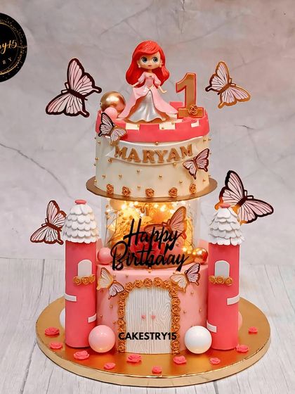 This two-tier chocolate castle cake is fit for a princess's first birthday. It features a detailed fondant castle design, a light-up spacer for a magical glow, and a charming princess toy topper.
