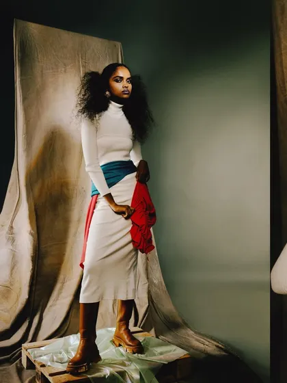 A full-length shot of 'ANAINA' in a color-blocked outfit. The pose is strong and statuesque, emphasizing the long lines of the skirt and the bold colors.