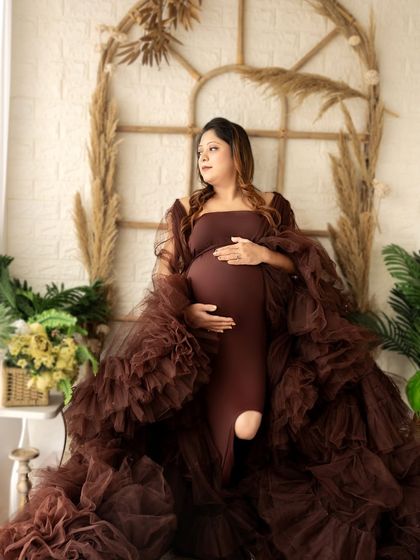 A classic maternity pose in a boho-chic setting. The expecting mother looks off to the side, framed by a rustic window arch and dried pampas grass.