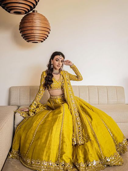 A full-length portrait of the bride seated on a sofa, her vibrant yellow lehenga spread out beautifully, showcasing its intricate mirror work.