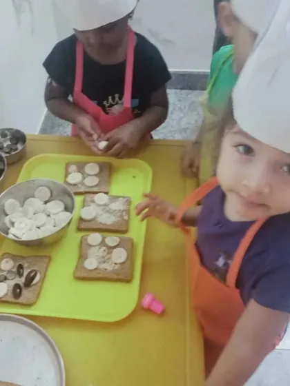 Our little chefs in their hats and aprons, preparing a healthy snack of toast with bananas and olives. Cooking activities teach children about nutrition, following instructions, and kitchen safety.