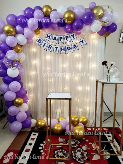 A beautiful purple, white, and gold balloon arch against a fairy-lit curtain. This setup is perfect for a simple yet elegant birthday celebration at home.