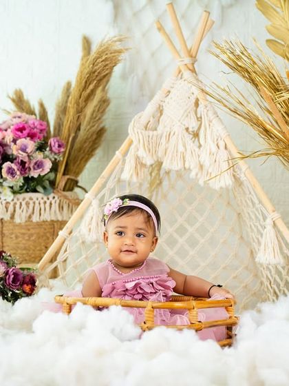 A lovely portrait of a toddler in a pink dress, sitting in a basket amidst clouds and flowers with a boho teepee in the background.