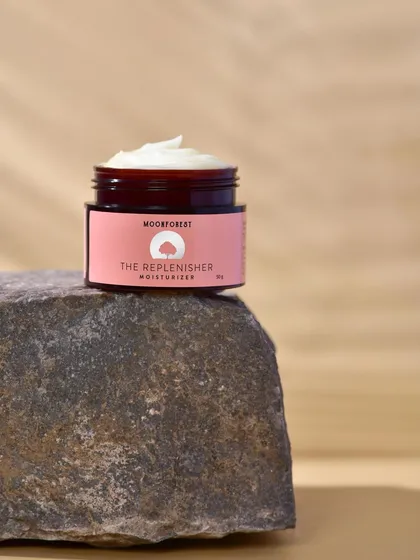 A close-up of "The Replenisher" moisturizer by Moon Forest, placed on a large stone. The contrast in textures between the smooth jar and the rough stone is visually interesting.