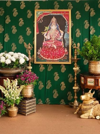 A traditional South Indian decor setup featuring a green paisley backdrop, a Tanjore painting, and an array of brass artifacts including a Nandi statue and flower vases.