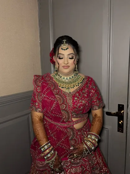 A soft and romantic bridal look. The makeup features a gentle smokey eye and a pink lip, perfectly complementing her red lehenga and green jewellery.