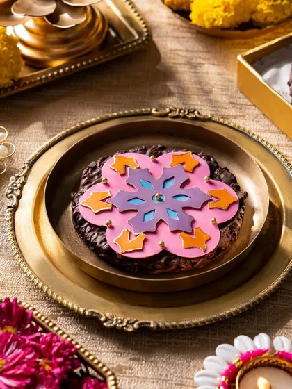 A large, dark chocolate cookie decorated with a vibrant, edible rangoli pattern, presented on a traditional brass plate. This is a unique and artistic treat that serves as a beautiful centerpiece for your Diwali celebrations.
