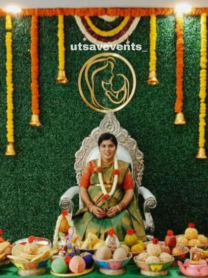 A photo of the mother-to-be at her baby shower, seated in front of a beautiful grass wall backdrop decorated with marigold garlands and hanging bells.