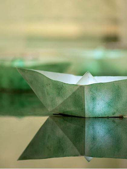 A detail from the 'Cherish' installation: a simple paper boat floats on a reflective surface. This small moment captures the theme of finding joy in simple, fleeting experiences.