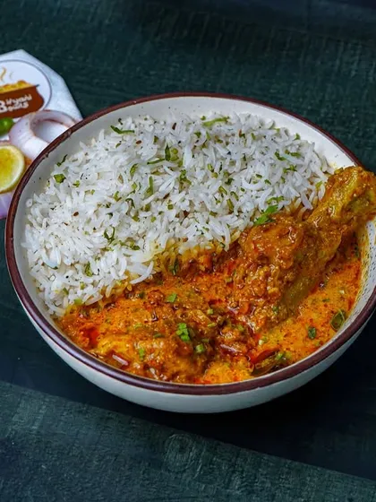 A bowl of chicken curry with a leg piece, served with a side of jeera rice. The dark, textured background makes the rich, orange gravy of the curry look even more appetizing.