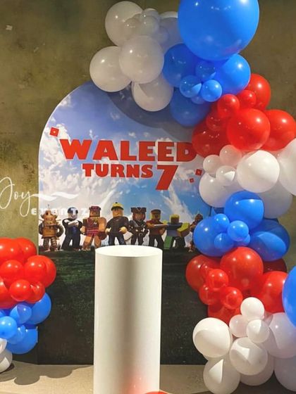 This Roblox themed setup for Waleed's 7th birthday uses a custom arched backdrop and a bold red, white, and blue balloon garland. A white plinth stands ready in the center for the birthday cake.