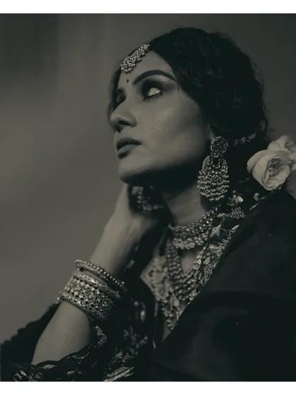 This black and white portrait showcases the side profile, highlighting the stacked bangles, elaborate jhumkas, and the soft texture of the rose in her hair. It's a study in traditional bridal adornment.