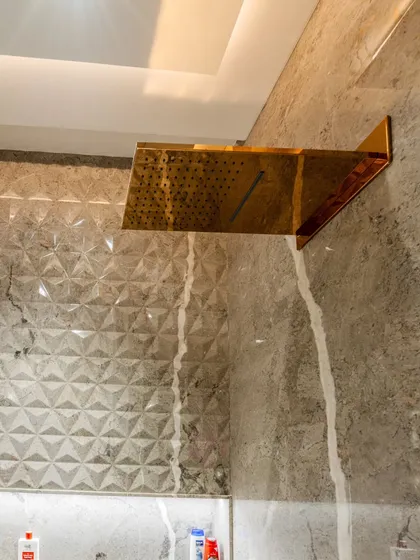 A close-up of the stunning gold-finished waterfall showerhead, set against a 3D geometric tile pattern. This detail adds a touch of unexpected luxury and texture.
