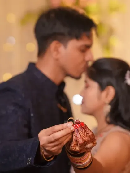 A close-up, artistic shot focusing on the engagement rings. The groom kisses the bride's forehead in the soft-focus background, creating a romantic and symbolic image.