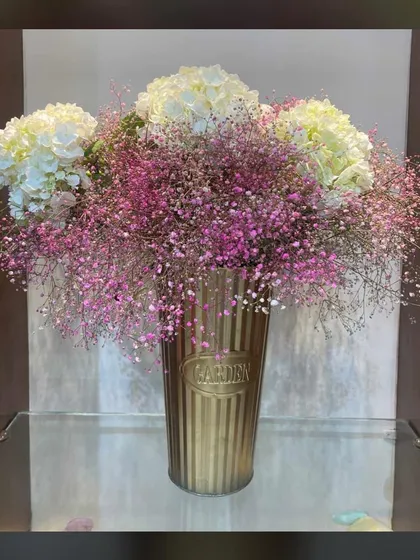 A beautiful vase arrangement featuring large white hydrangeas and delicate pink and white baby's breath. The golden "Garden" vase adds a touch of vintage charm.