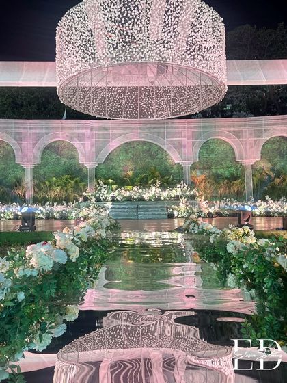 The aisle, lined with abundant white flowers and greenery, leads to a magnificent stage. The reflection of the massive baby's breath chandelier on the mirrored walkway creates a stunning, dreamlike effect.