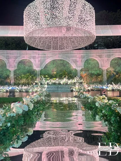 A stunning reflective aisle mirrors the grand, illuminated floral chandelier above, creating a magical pathway for the couple. The aisle is lined with lush white roses and greenery, leading to a stage set against a wall of living plants.