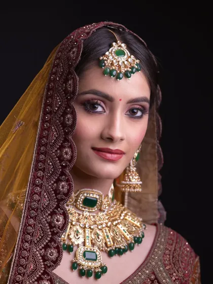 A close-up of a traditional bridal look with heavy green and gold jewelry. The makeup is bold and defined.