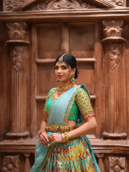 A portrait of a model in a designer half saree, smiling brightly. The intricate wooden door in the background adds a touch of traditional architecture to the shot.