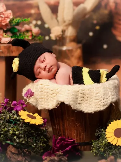 A newborn in a bee costume sleeps in a bucket filled with soft fabric, surrounded by sunflowers and other floral elements for a garden feel.