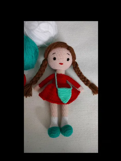 A handmade crochet doll with two long braids, a red dress, and a little green bag.
