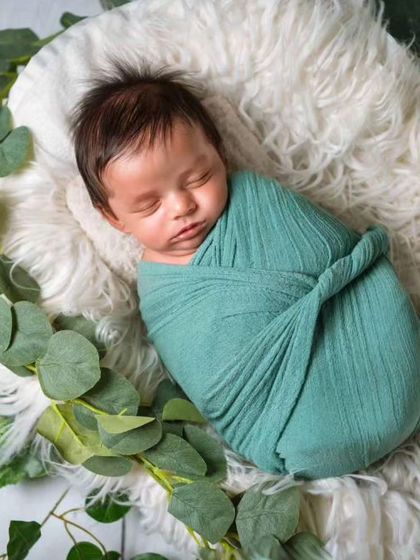 A newborn swaddled in green, resting on a white fur rug surrounded by eucalyptus leaves for a fresh, natural look.