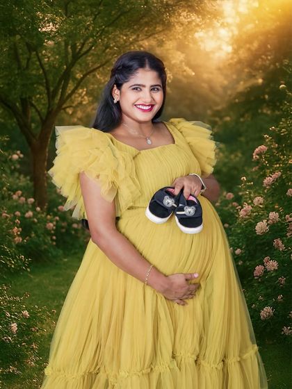 A radiant outdoor portrait of a mother-to-be in a lovely yellow gown, holding a pair of baby shoes. The golden light and garden setting create a warm, dreamy atmosphere.