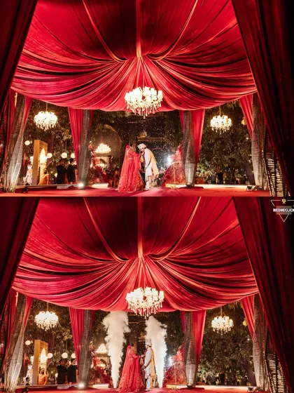 A dramatic wide shot of the Varmala ceremony, framed by rich red drapery and magnificent chandeliers, highlighting the grandeur of the wedding decor.