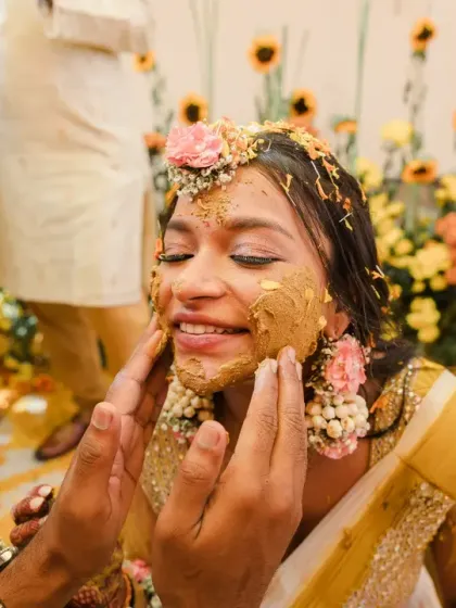 A close-up of the bride's smiling face as she gets haldi applied. The vibrant yellow paste and flower jewellery create a beautiful, traditional Haldi portrait.