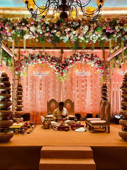 A grand indoor mandap setup with a blush pink theme, featuring a ceiling of cascading flowers and elegant traditional seating for the ceremony.