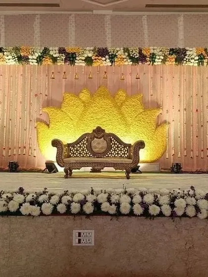 A lovely reception stage with a large, lotus-shaped backdrop made of yellow flowers. The stage is framed with a mix of colorful flowers and a curtain of delicate white flower strings.