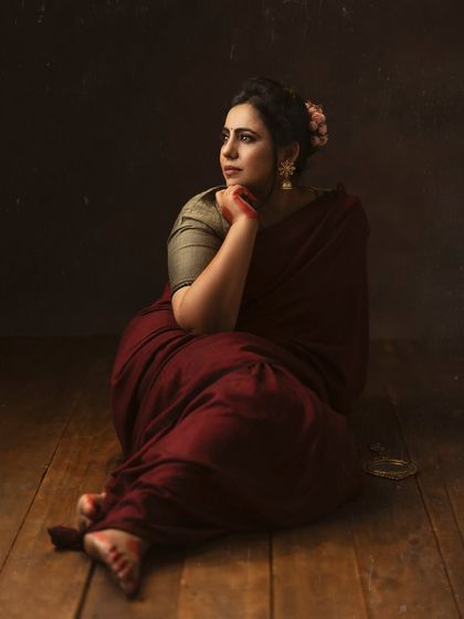 Elegance personified. A woman in a deep red saree, seated on the floor in a moment of quiet contemplation. The wooden floorboards add warmth and texture.