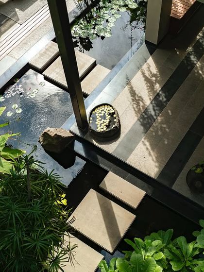 An aerial view of a courtyard with a dark water body, crossed by stone steps. The water reflects the sky and surrounding plants, adding depth and a sense of calm to the geometric layout.