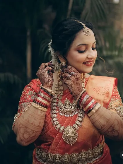 A classic bridal pose, adjusting an earring, which highlights the intricate temple jewellery and mehndi.