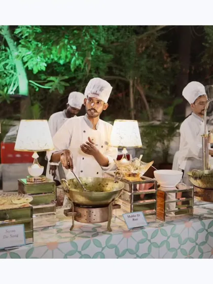 A chef at a live counter preparing a traditional Indian curry in a brass kadai, surrounded by fresh ingredients and elegant serveware.