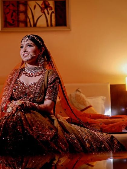 A wide shot of a bride sitting on a bed, her beautiful lehenga spread out on the reflective floor.