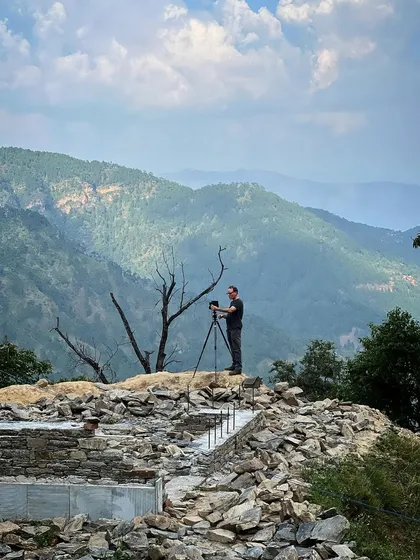 A photographer stands atop a pile of stones to document the construction progress. The dramatic landscape of the Himalayas provides an unparalleled backdrop for capturing the story of our architecture.
