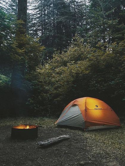 A solitary tent glows from within, nestled in a dense forest clearing. The nearby fire pit promises a warm, cozy evening under the canopy of trees. This is perfect for those seeking quiet and solitude.